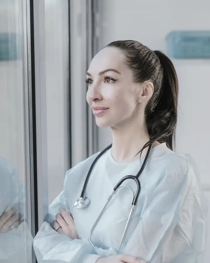 A surgeon in a pale gown with a stethoscope, looking out of a window