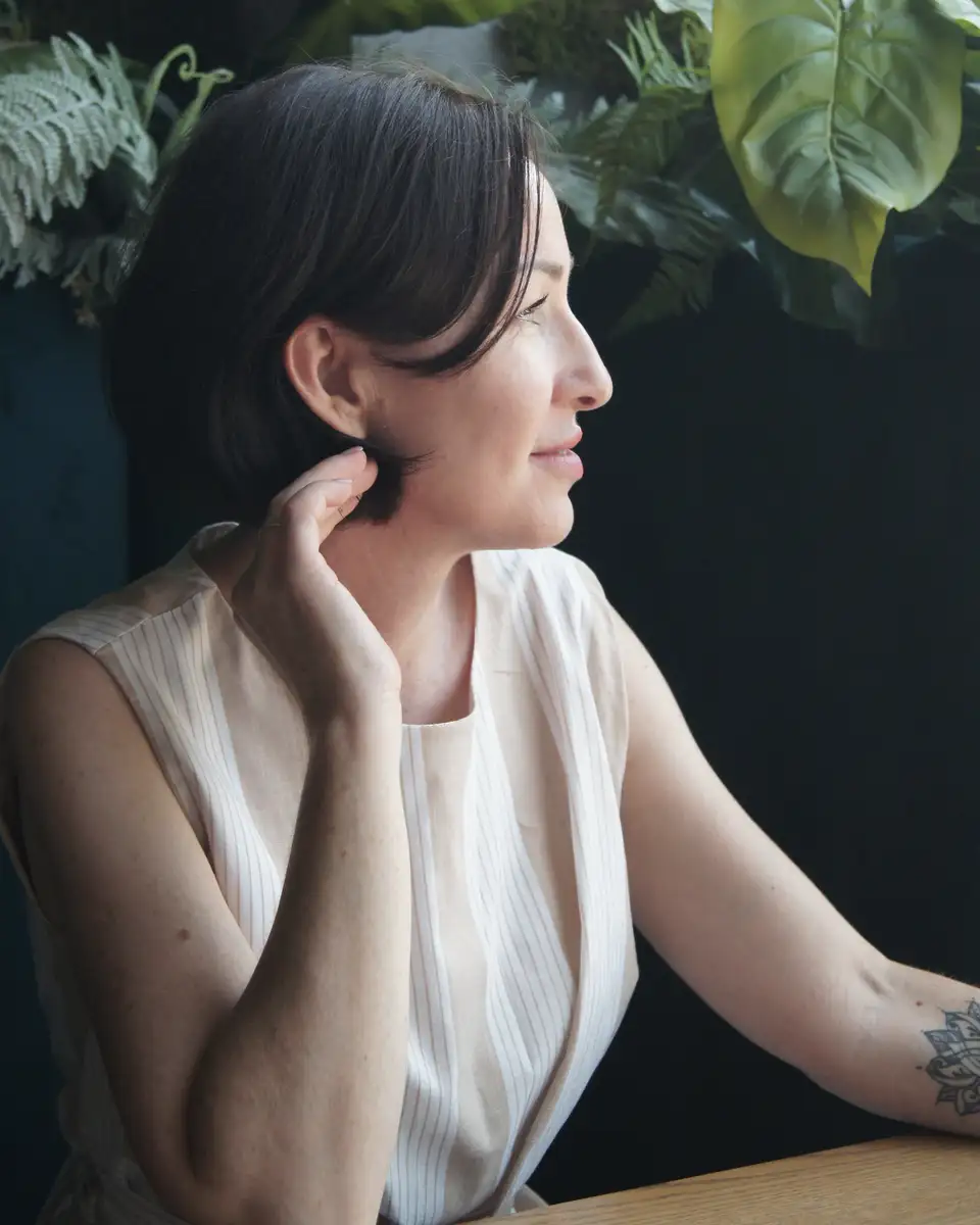 A woman with dark hair sitting by a window with plants, looking out thoughtfully