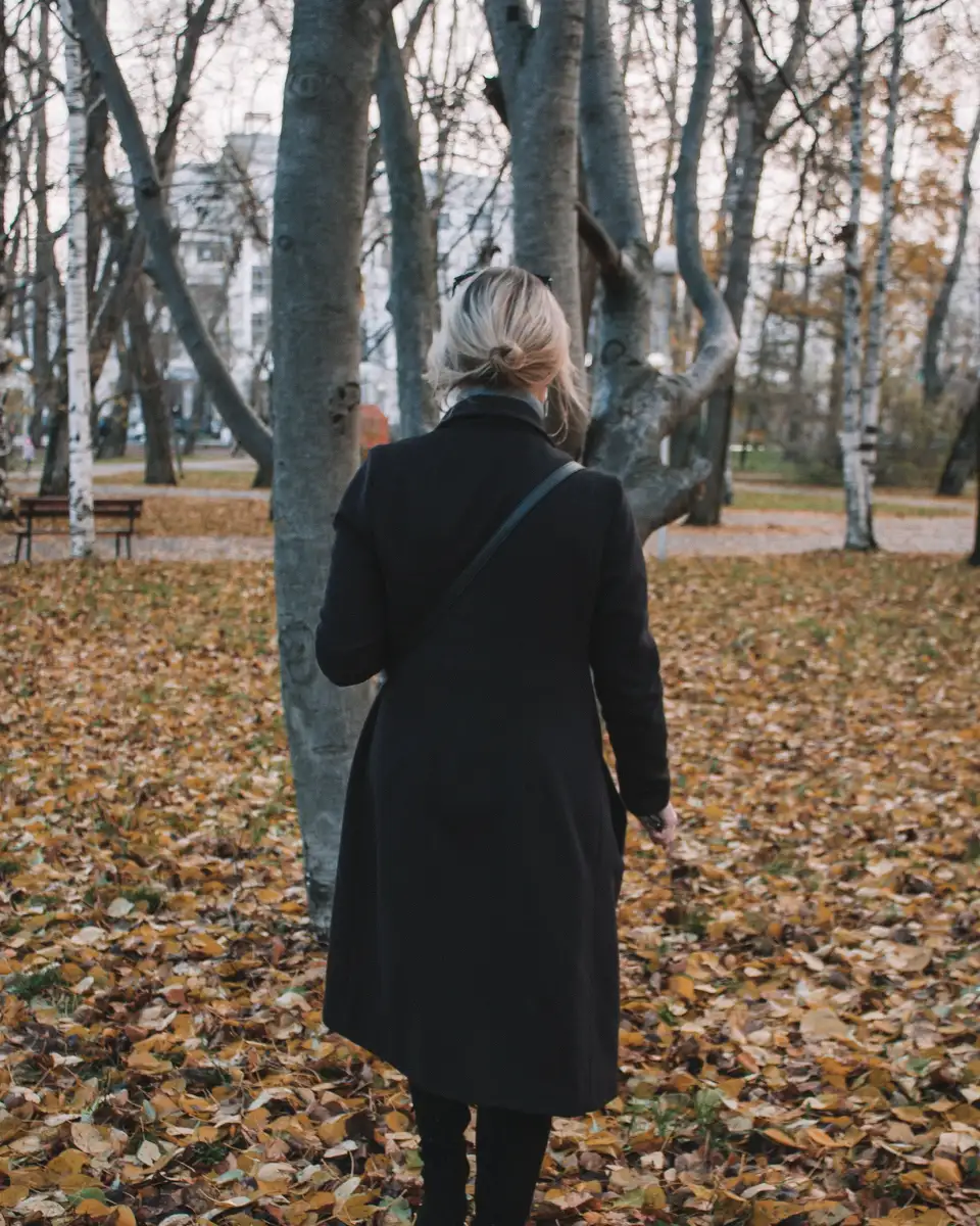 A woman in a dark coat walking away through a park of fallen leaves