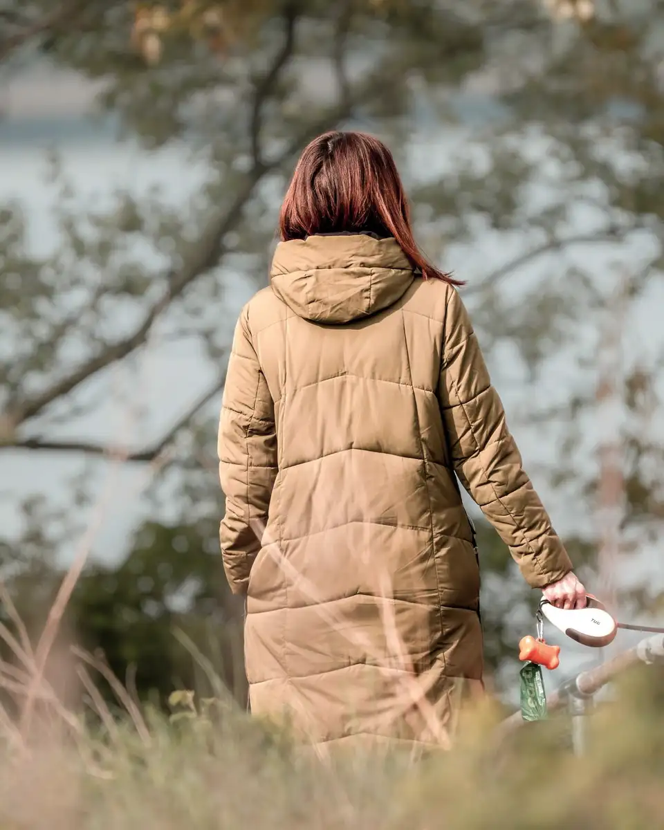 A woman in a camel coat walking away along a path by a lake in autumn